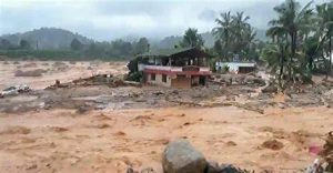 Mundakkai in Wayanad after the July 2024 landslide, with muddy floodwater covering roads and houses among palm trees under cloudy skies.